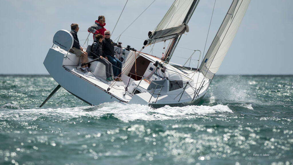 Segelboot mit Crew auf Deck, seitlich gekippt, Wellen spritzen.